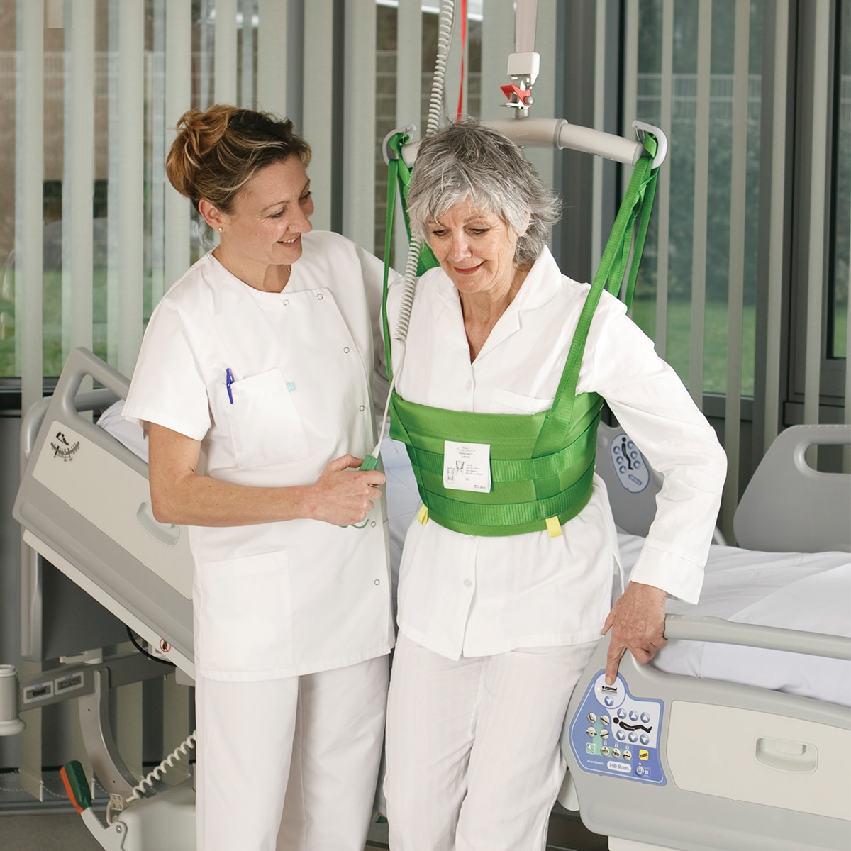 Un clinicien aide un patient à marcher dans une chambre d’hôpital avec lève-personne plafonnier de Hillrom et un dispositif MasterVest.